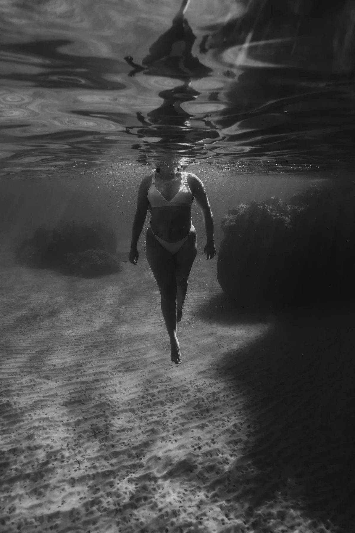 filmische onderwater foto van een vrouw in zwart-wit, ze beweegt naar het oppervlak, haar lichaam is in een elegante beweging vastgelegd. Ze draagt een lichte bikini.