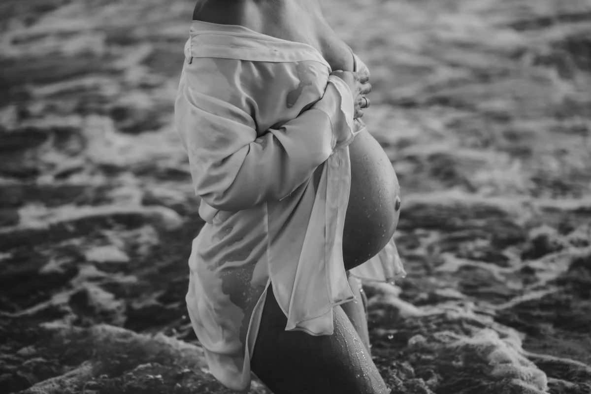 Een moderne boudoir foto van een zwangere vrouw in een openvallende oversized blazer op het strand. Het contrast tussen de strakke lijnen van de jas en de zachte vormen van haar lichaam wordt versterkt door harde schaduwen.