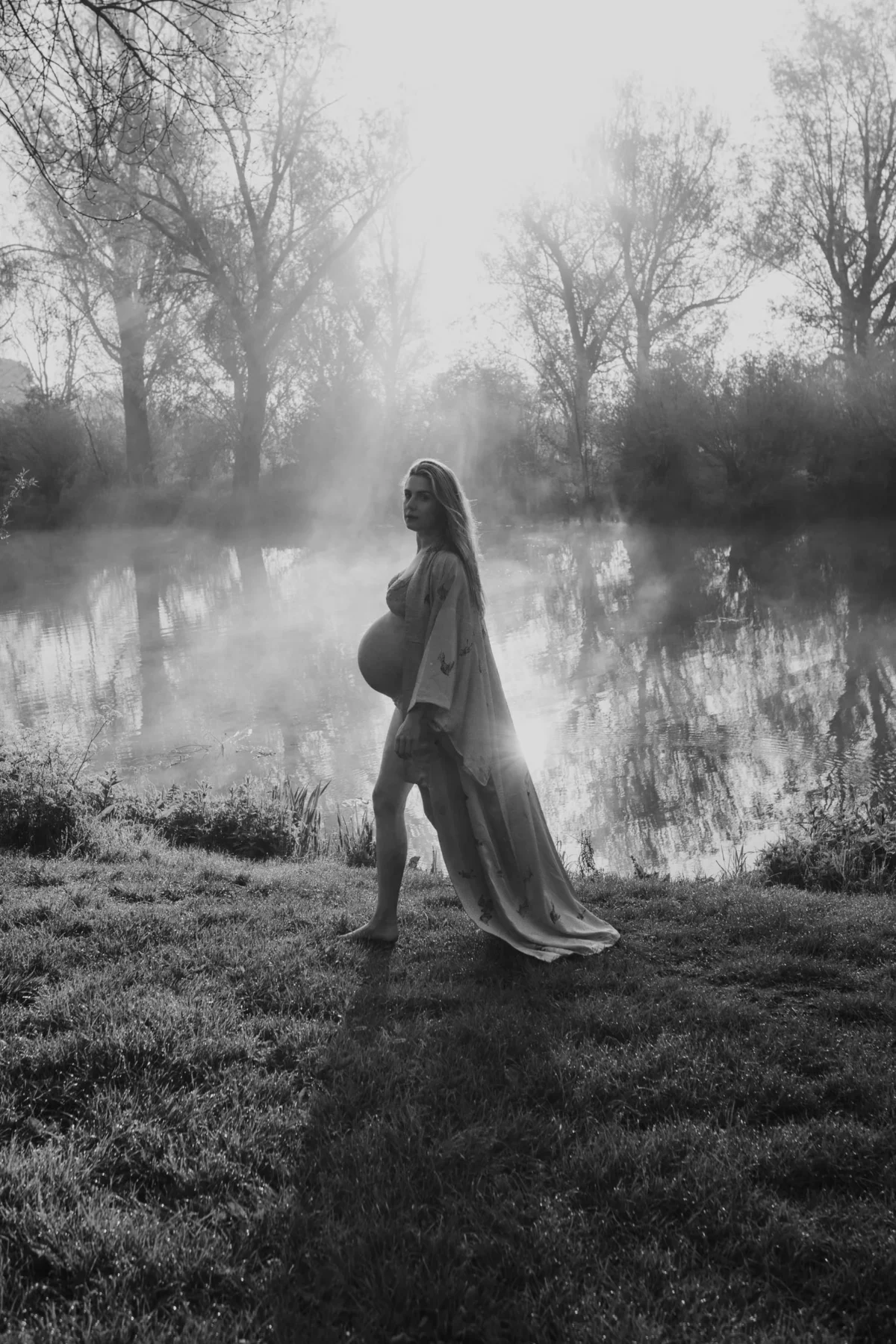 dromerige zwangerschaps fotoshoot van vrouw die langs een meer loopt in de buitenlucht met mist en bomen om het meer heen.