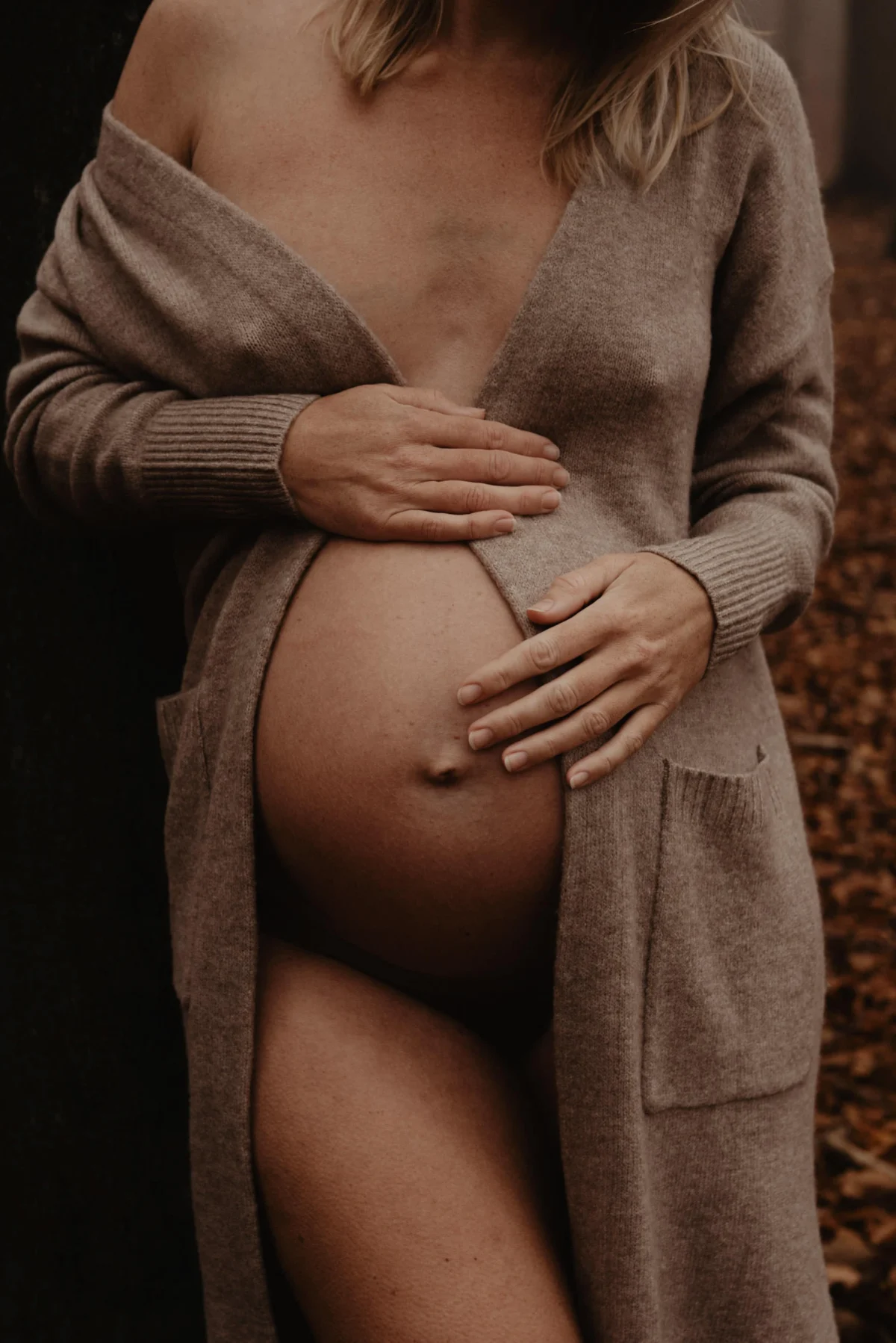 Zwangere vrouw met lange zandkleurige kimono die open slaat om de buik met hand op haar buik.