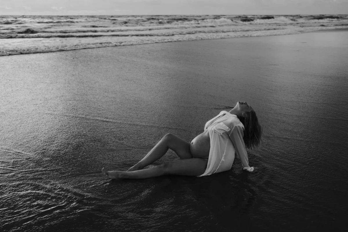 dromerige zwangereschapsfoto van vrouw die naar achteren leunt met holle rug waardoor witte blouse open slaat en de buik laat zien, ze ligt aan het strand naast de branding.