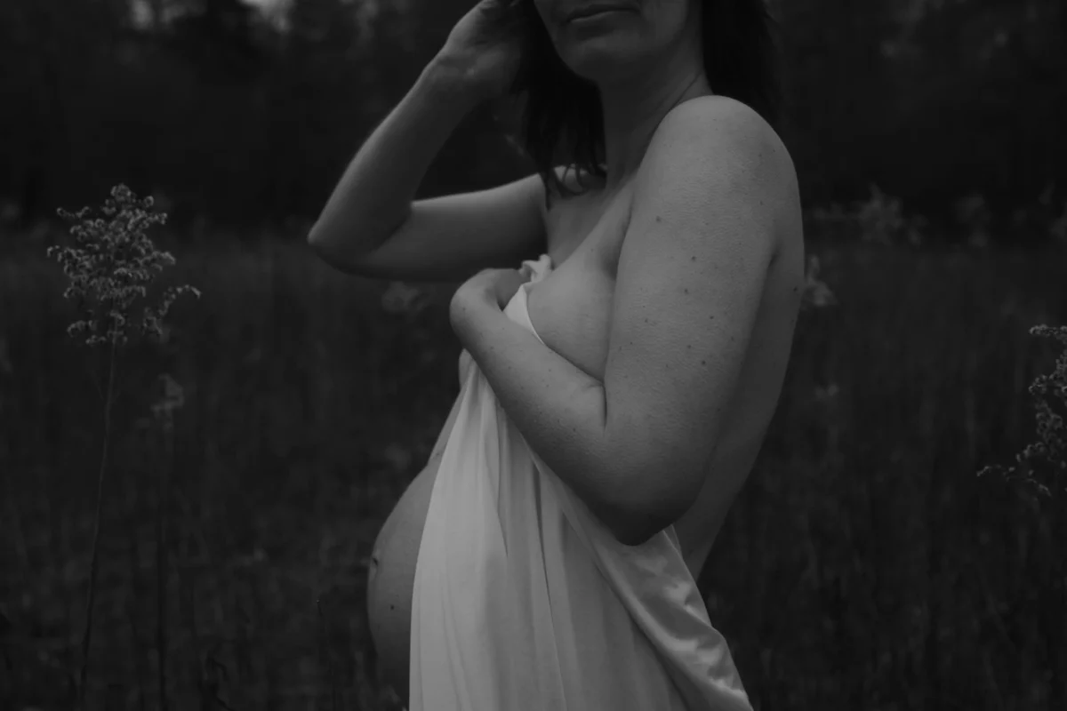 contrast foto van donkere achtergond en voorgrond een zwangere vrouw met lichte huid en wit laken die haar borsten en buik deels verhullen maar niet genoeg om niet te kunnen zien dat zij zwanger is.