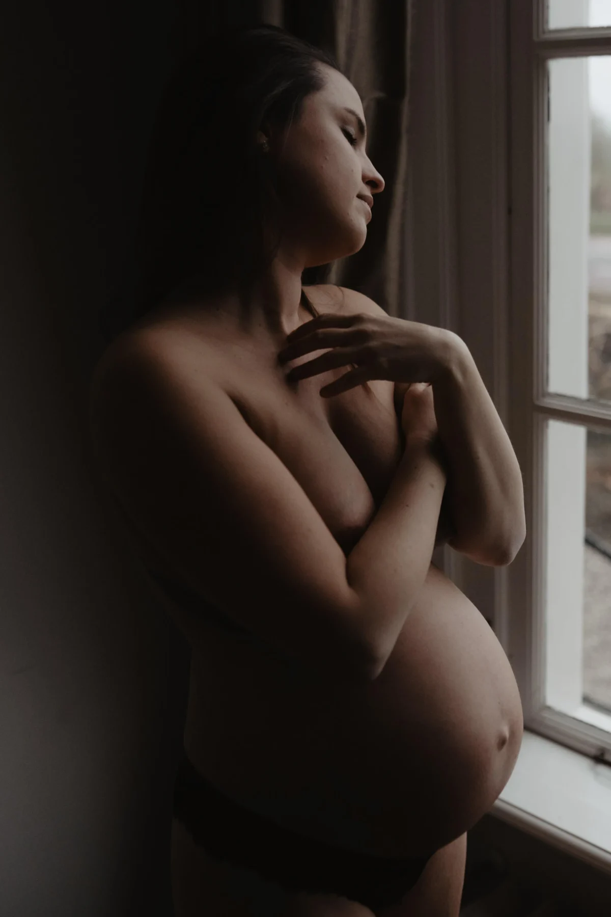 zwangere vrouw staat voor een raam waardoor een natuurlijk licht opp haar buik en hals en schouder schijnt terwijl de rest van haar lichaam in schaduw gehuld is voor een dromerig effect, speels licht voor je zwangerschapsshoot van Shapes Shadows.