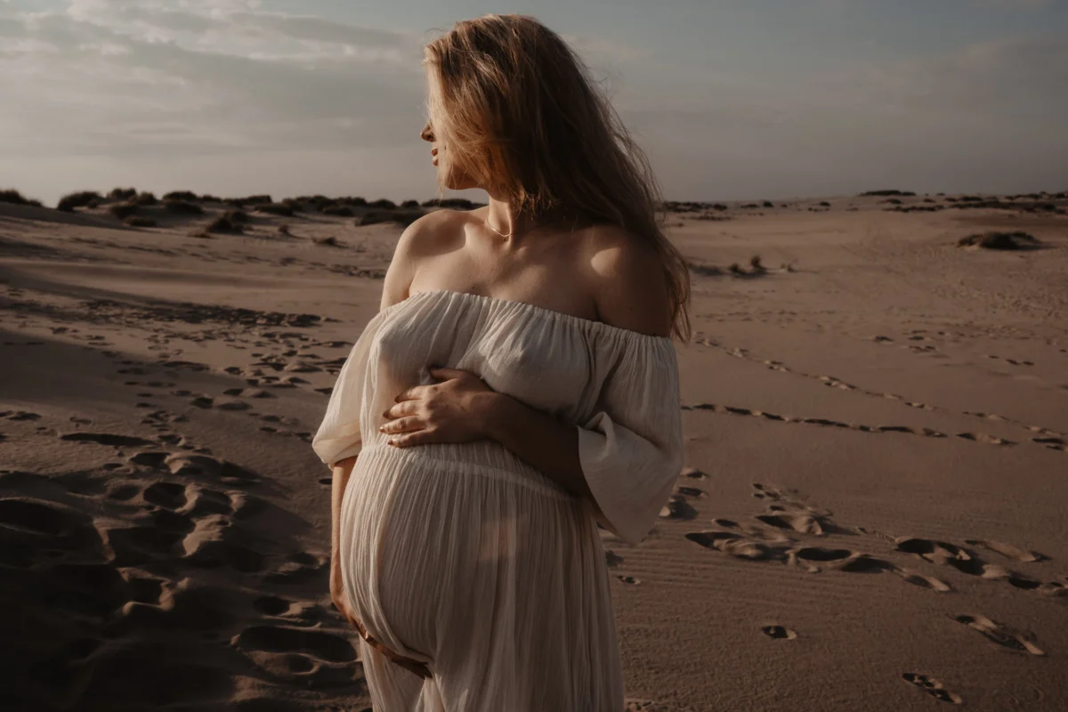 elegant portret van zwangere vrouw die haar hoofd weg draait van de camera, haar ronde buik en borsten spieken door de witte jurk heen die losjes over haar lichaam hangt in het avondzon licht.