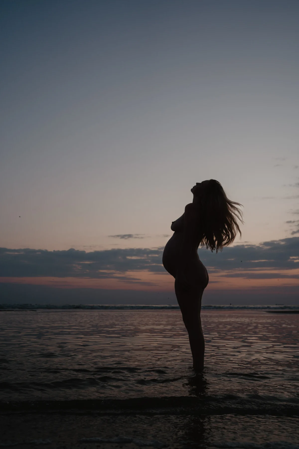 Silhouette van zwangere vrouw achterover buigend met haar lichaam aan het strand voor ondergaande zon, paarse en roze kleuren, natuurlijke kracht.