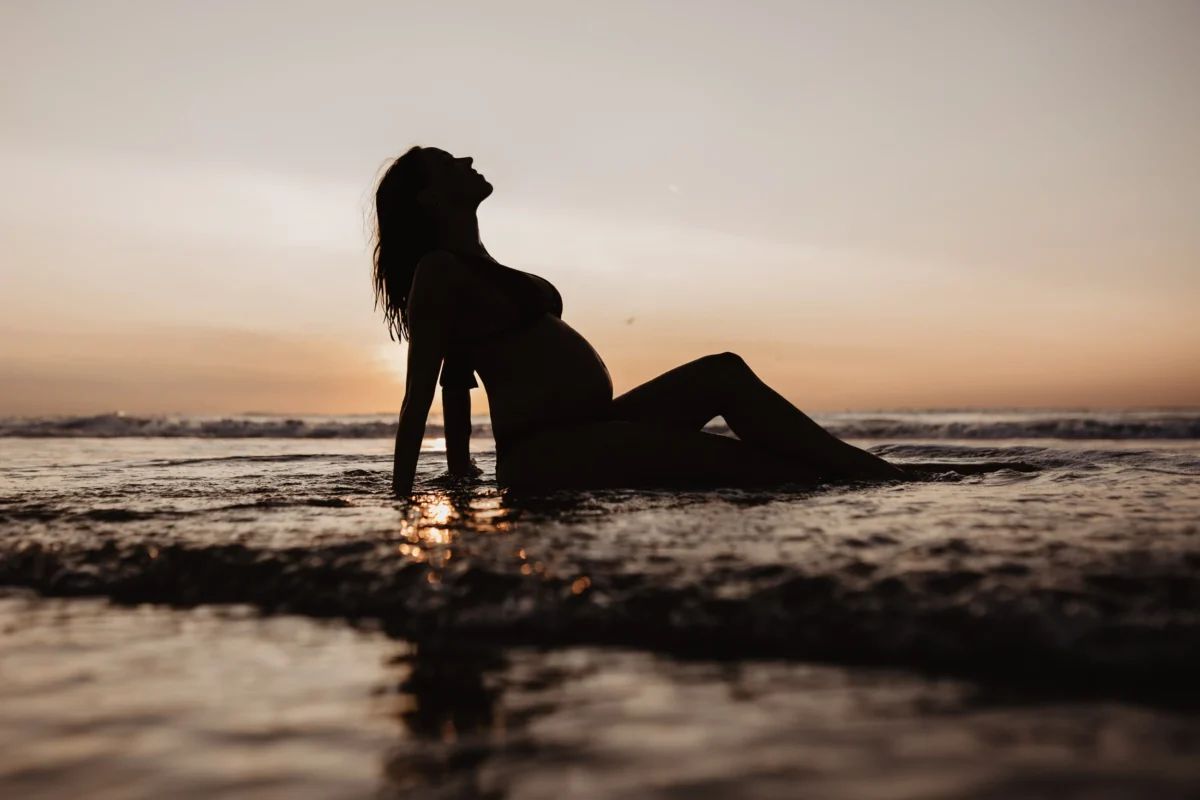 silhouette van zwangere vrouw di in de branding achterover leunt voor een bijna ondergaande zon.