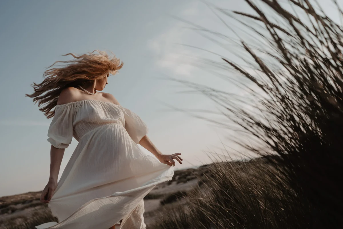 Zwangere vrouw in witte jurkt draait haar lichaam weg van de camera waardoor er een tijdloos vintage effect ontstaan, natuurlijke kleuren door de grassprieten die ook in beeld zijn,