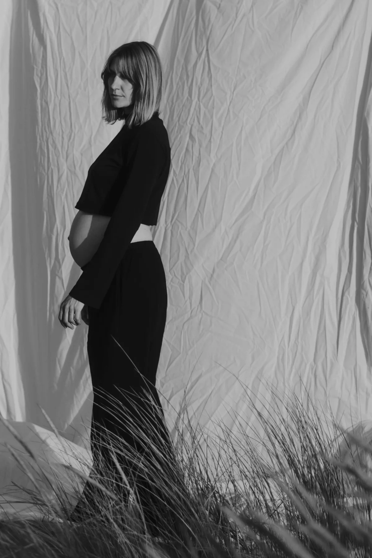 chique geklede zwangere vrouw poseert voor een wit laken buiten in het gras. Kwetsbaarheid en zelfverzekerdheid in contrast met elkaar. Zwart-wit portret door Shapes Shadows