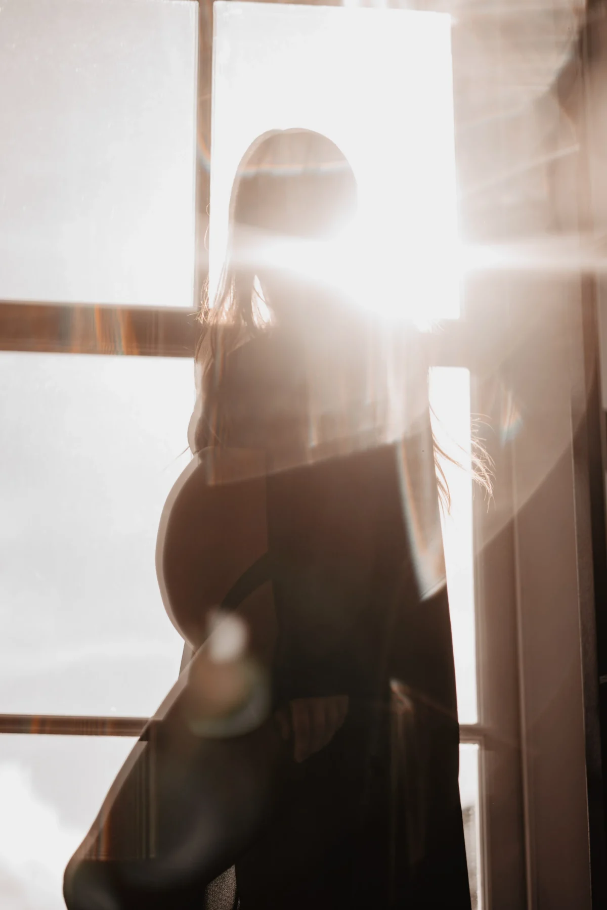 zon als lichtbron door raam overgiet het lichaam van een zwangere vrouw wardoor er een vintage effect ontstaat en de vrouw geheel in schaduw gehuld is en haar contouren scherp afsteken tegen de zonovergoten ramen