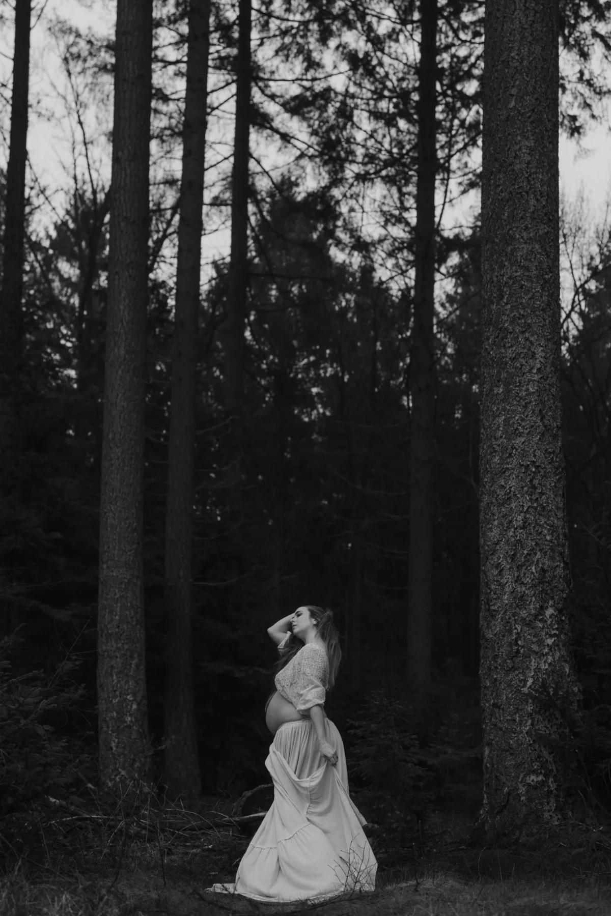 zwangere vrouw met wit doek over lichaam poserend tussen hoge bomen, zart-wit shapes shadows zwangerschapsfotografie, tijdloos en artistiek