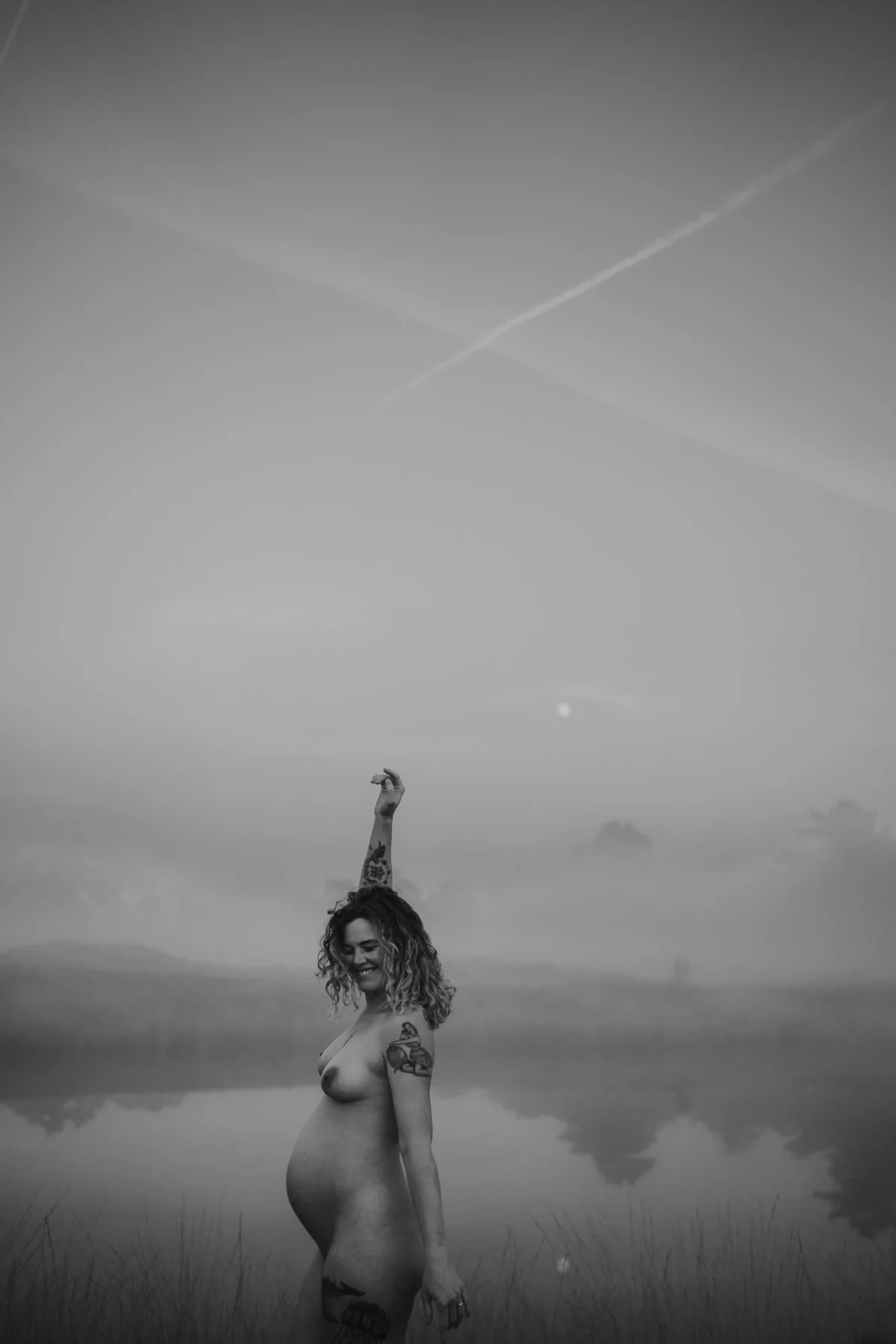 happy zwangerschaps portret te zien aan pose van vrouw, een arm omhoog, naakt ind e natuur voor een plas met water.
