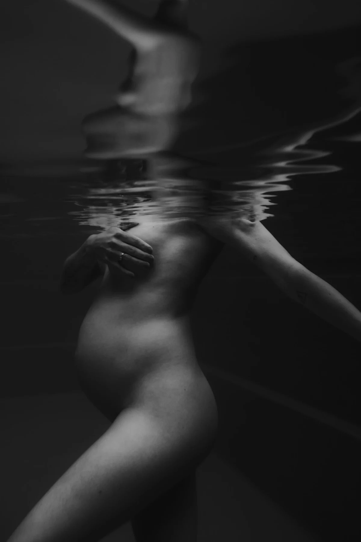 onderwater zwangerschapsfoto in zwart-wit vrouw te zien van zijkant en bedekt haar borst met hand, hoofd in in de schaduw gehuld