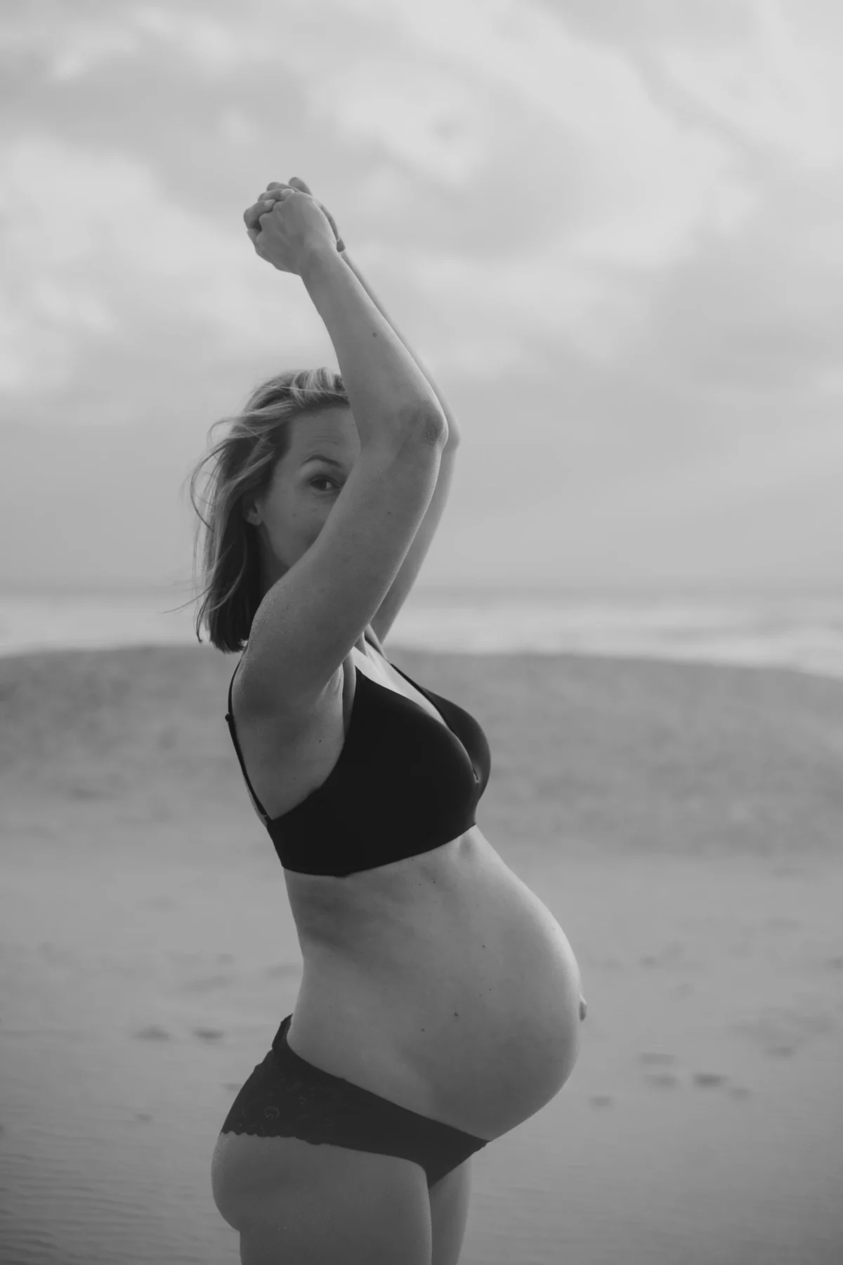 Zwart-wit foto van zijkant van zwangere vrouw die haar armen omhoog heeft zodat deels haar gezicht verborgen blijft, ze is op het strand