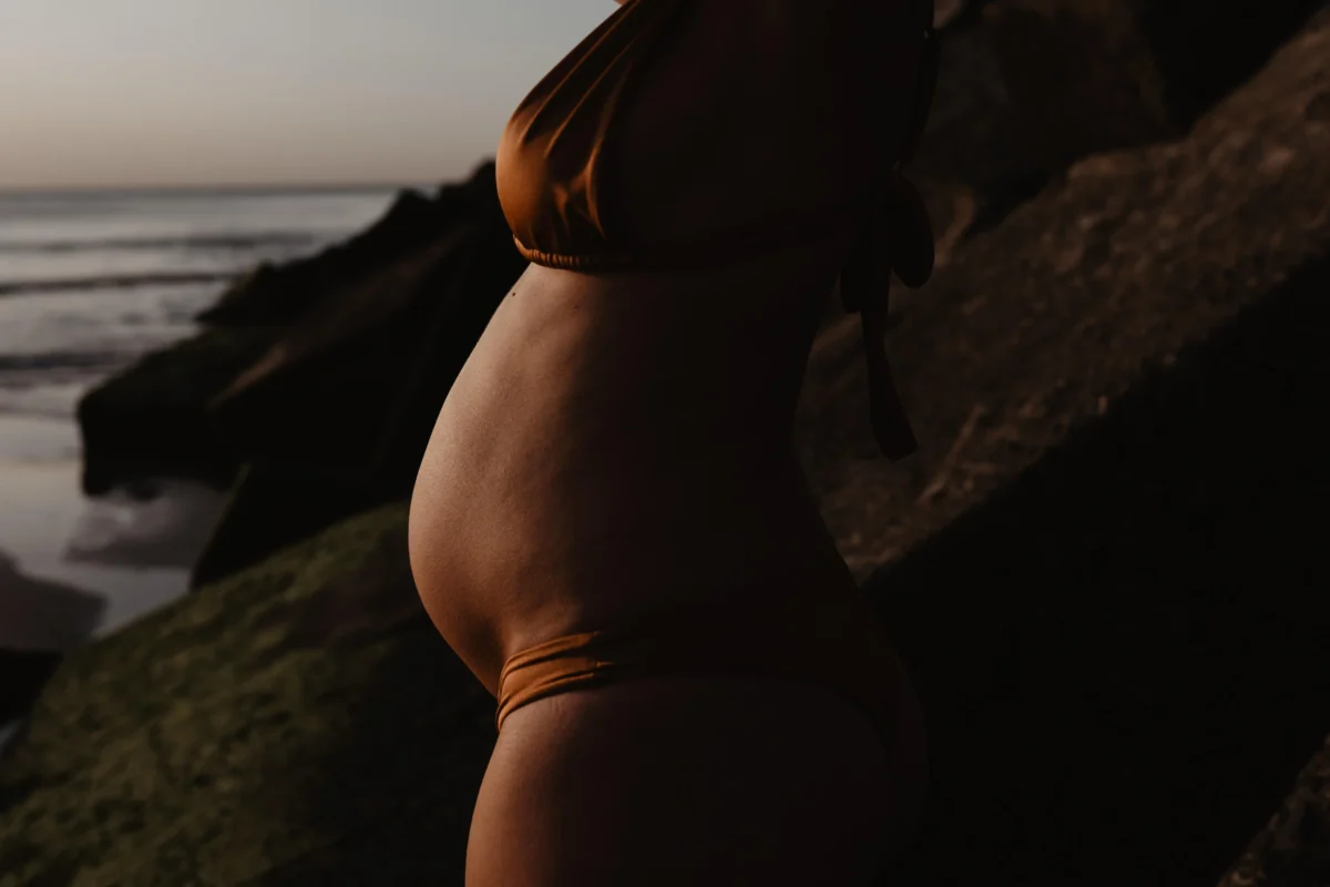 een ondergaande zon zorgt voor een koper effect van de huid van een zwangere vrouw met een oranje bikini, de buik is in focus, je ziet de zijkant van het lichaam