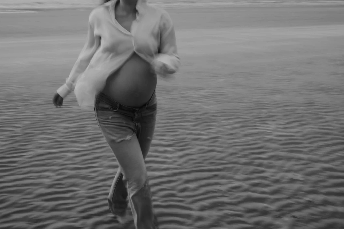 Een moderne boudoirfoto van een zwangere vrouw in een openvallende oversized blouse. Het contrast tussen de strakke lijnen van de jas en de zachte vormen van haar lichaam wordt versterkt door harde schaduwen.