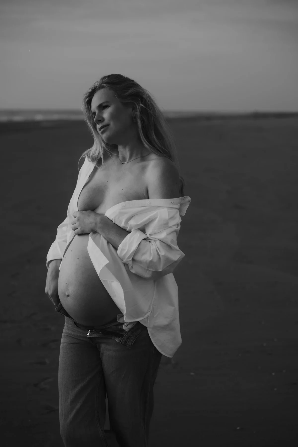 klassieke zwangerschaps fotografie van vrouw met oversized witte blouse waar buik doorheen komt op het strand met hoofd een beetje getilt en natuurlijke uitstraling.