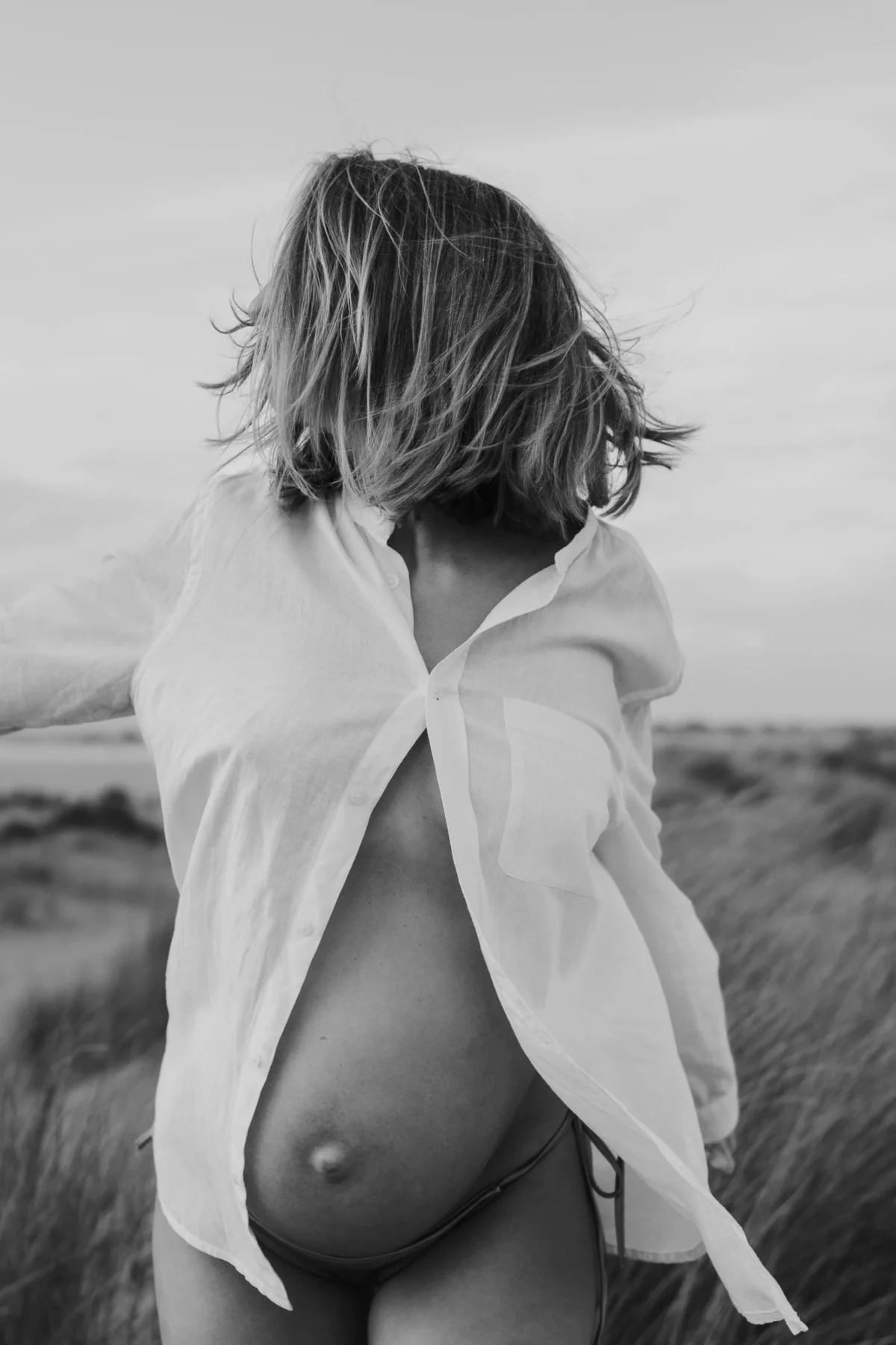 klassieke zwangerschaps fotografie van vrouw met oversized witte blouse waar buik doorheen komt aan het strand met haar voor gezicht bewogen.