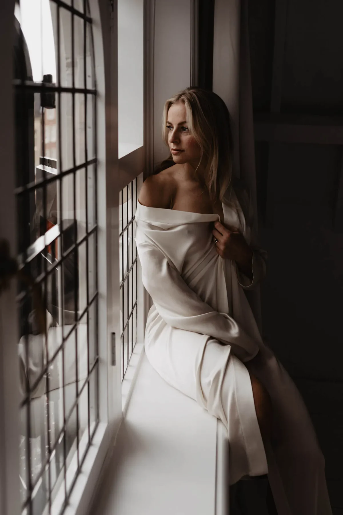 vrouw zit op stoel voor raam in wit laken gehuld en kijkt dromerig uit het raam