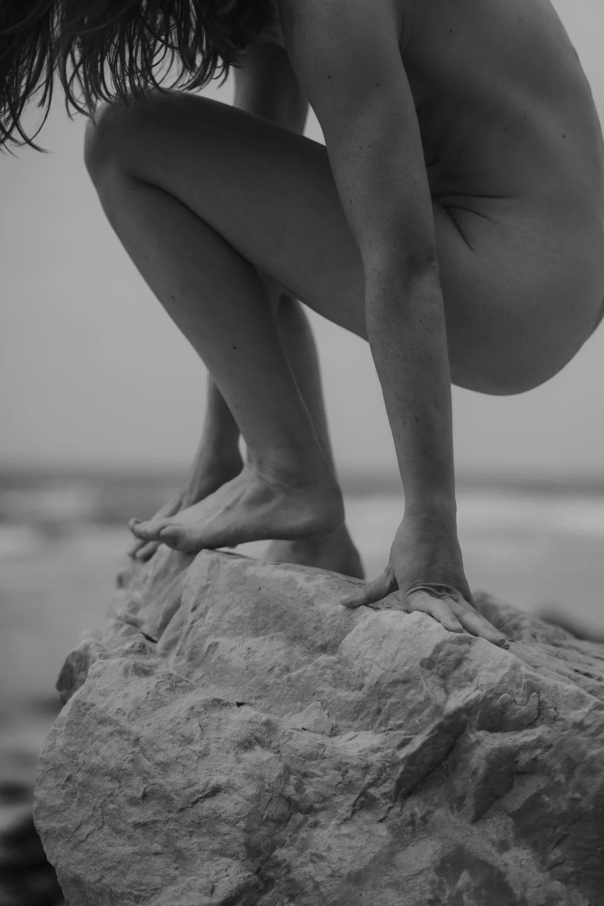 vrouw staat in een zelfverzekerde pose tijdens haar boudoir shot op een witte rots op een ruig desolaat Ibiza strand, close up van handen, armen, benen en voeten in hurk stand.