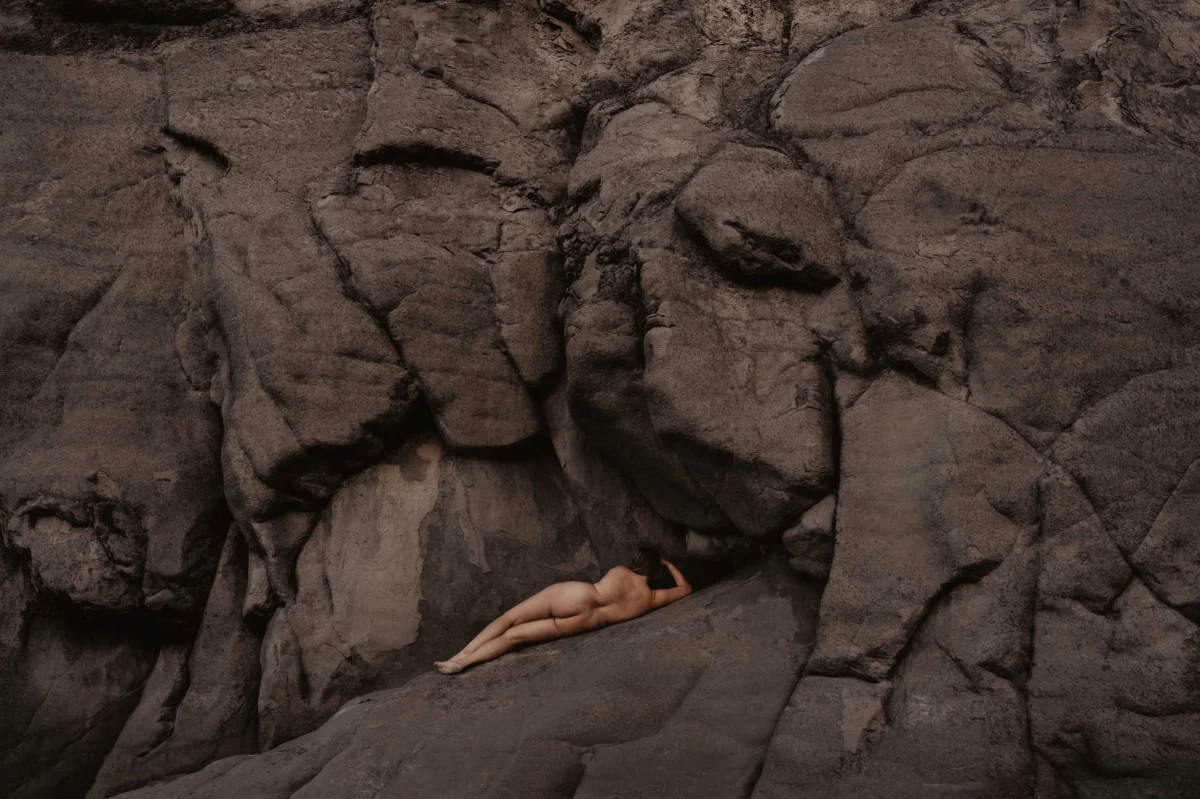 donkere rotspartij met naakte vrouw die in contrast maar ook eenheid is met de ruigheid van haar omgeving, haar kleur is warm en ze ligt er relaxed bij, ze is van achteren te zien.