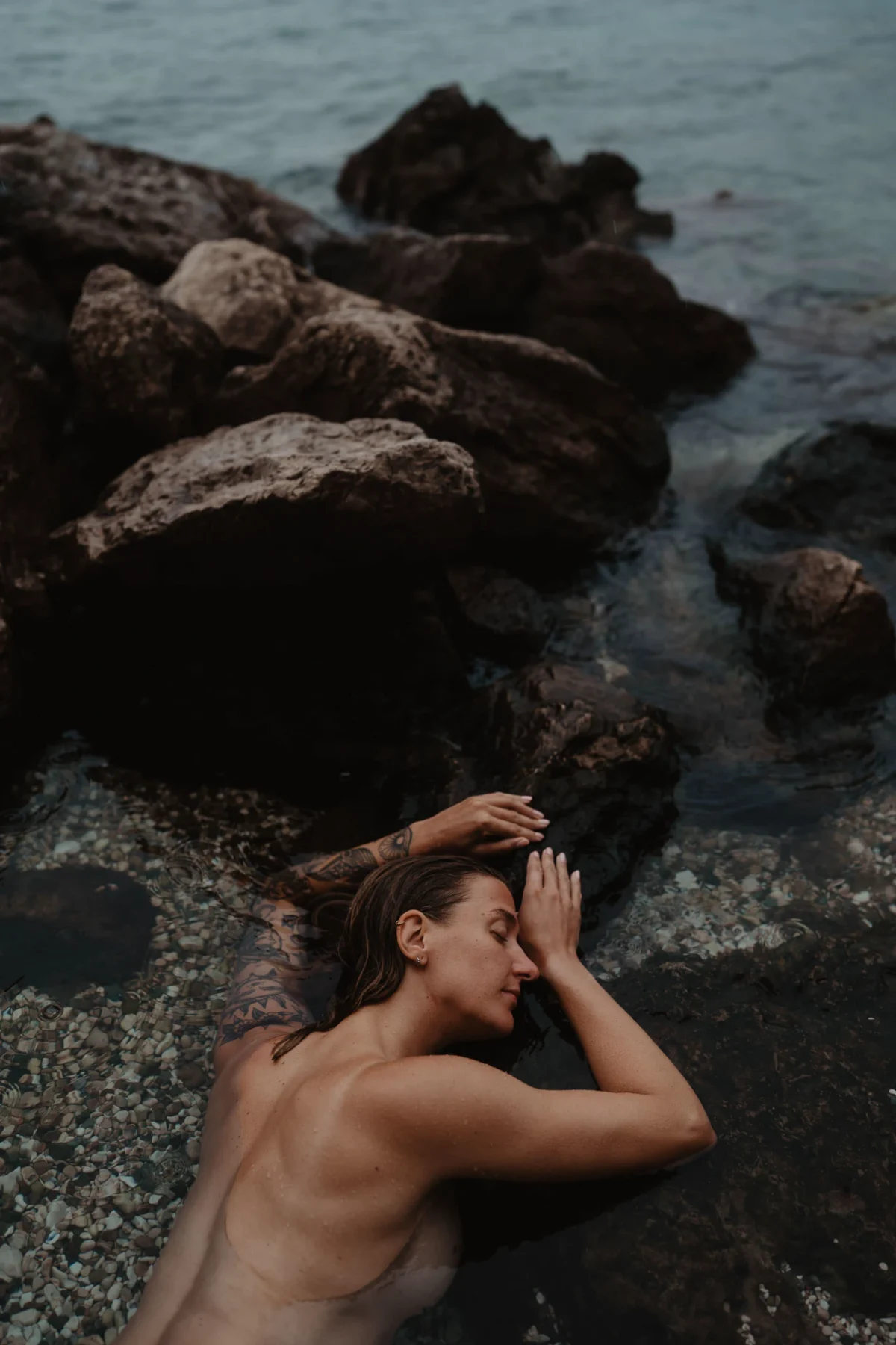dromerige foto van een vrouw die rust op een rots en half in het water licht als een zeemeermin, in warme tinten van de rots en kiezels gehuld.
