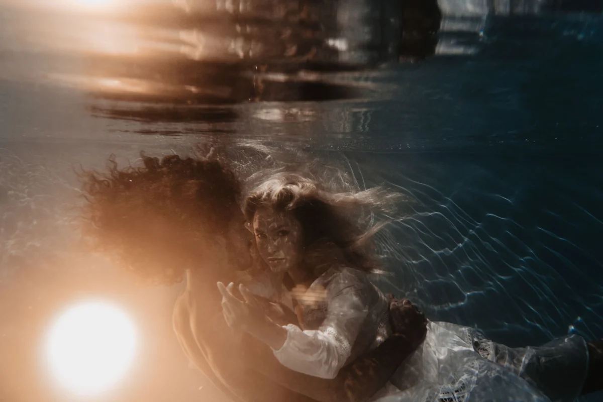 koppel onderwater waarvan de vrouw recht in de kamera kijkt, beiden hebben lang haar dat dromerig om hen geen drijft en de lichtbron komt van onderen, de vrouw draagt witte kleding. Alles is een sprookjesachtig geheel