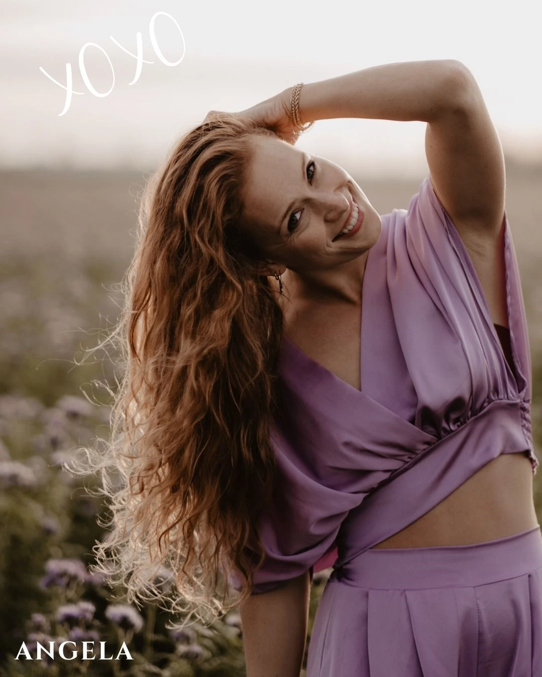 portret van boudoir fotograaf in Nederland Angela, buiten in een veld met bloemen met een lila top en broek aan, ze heeft haar rode haar vast en haar hoofd gekanteld naar links. Angela laat je van je beste kant zien en geeft je zelfvertrouwen voor je boudoir shoot