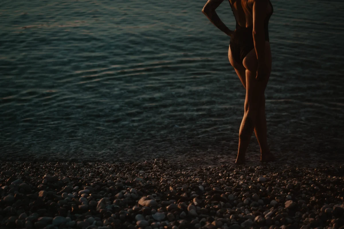 mystieke foto van vrouw op kiezelstrand met ondergaande avondzon die haar huid bronze en zacht kleurt. Alleen de billen en benen zijn te zien. De vrouw draagt een zwart badpak.