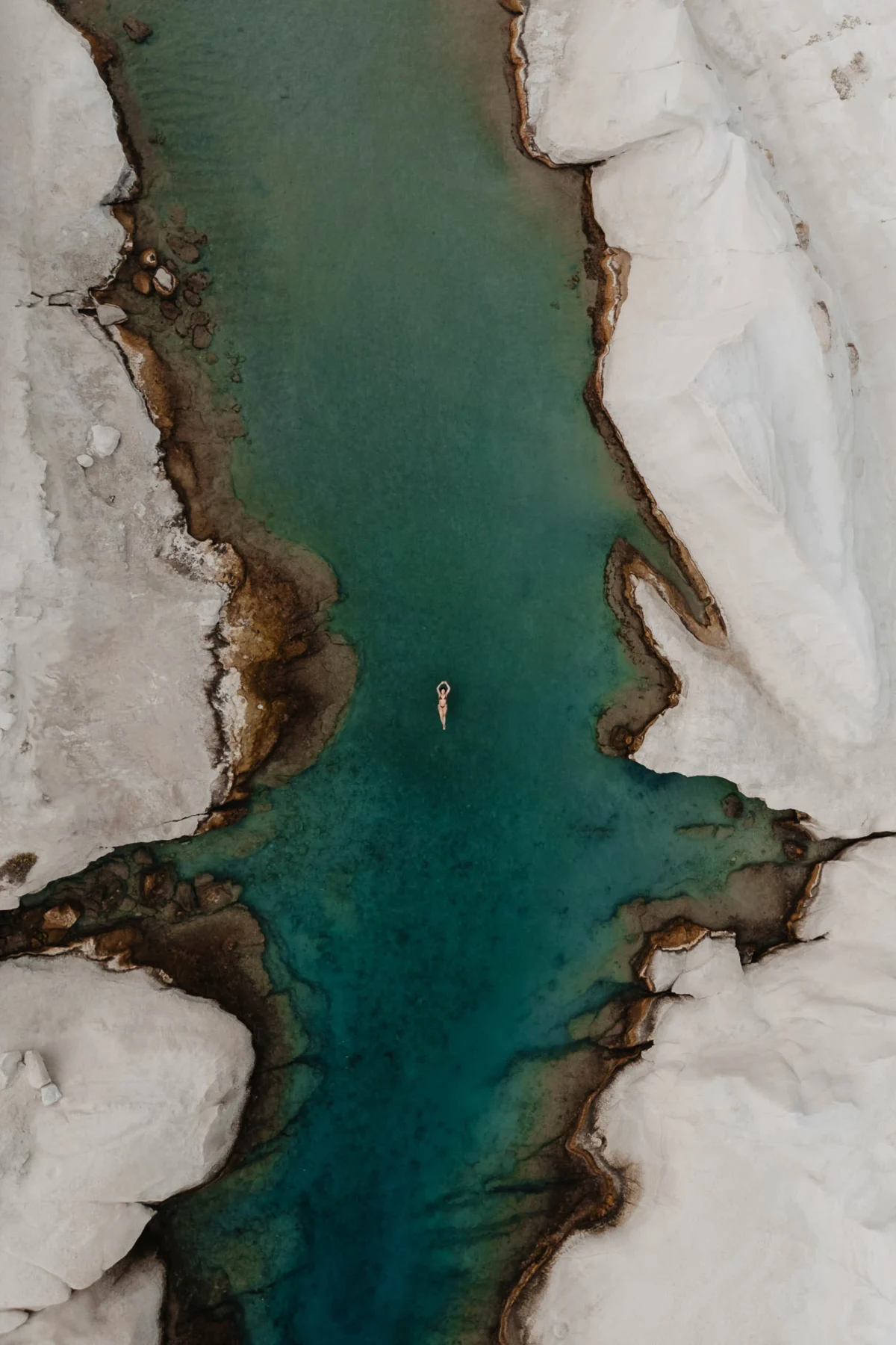 tijdloos beeld van een vrouw die van bovenaf met een drone gefotografeerd is liggend in het water tussen twee witte rotsen in. Het water is turquoise en waar de rots het water raakt is bruin. Het is een schitterende combinatie van ruwe natuur en menselijk schoon.