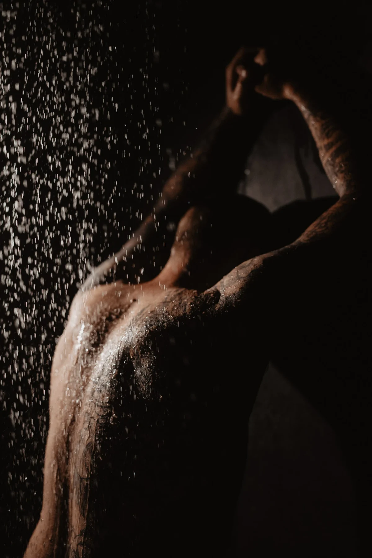 Man leunt tegen muur onder de douche tegen vrouw aan waardoor water op zijn rug spattert en de vrouw slechts een topje van het hoofd te zien is, liefde en kracht in dit koppel boudoir portret.