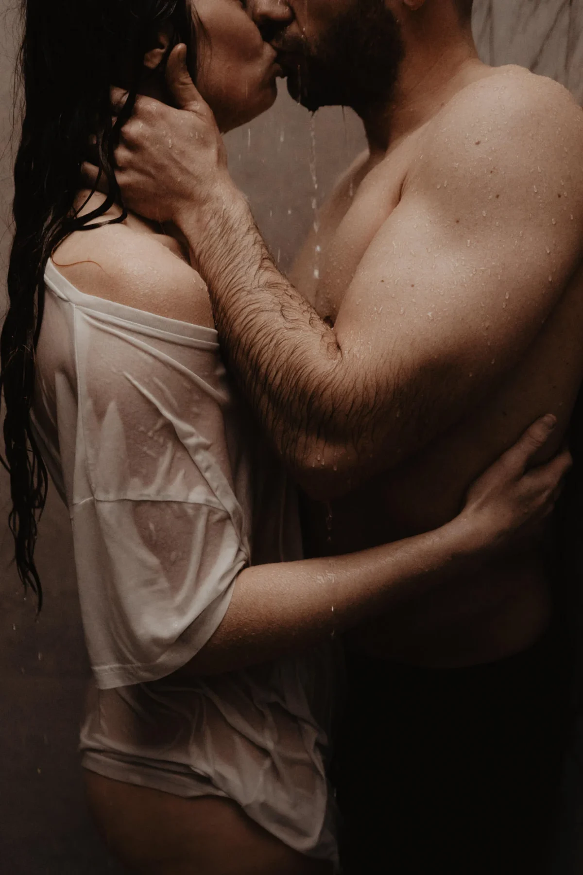 Man en vrouw onder de douche, vrouw met wit nat tshirt, alleen onderkaken van koppel te zien, man heeft met zijn gespierde armen gezicht van zijn partner zachtjes vast. Lief en warm boudoir portret.