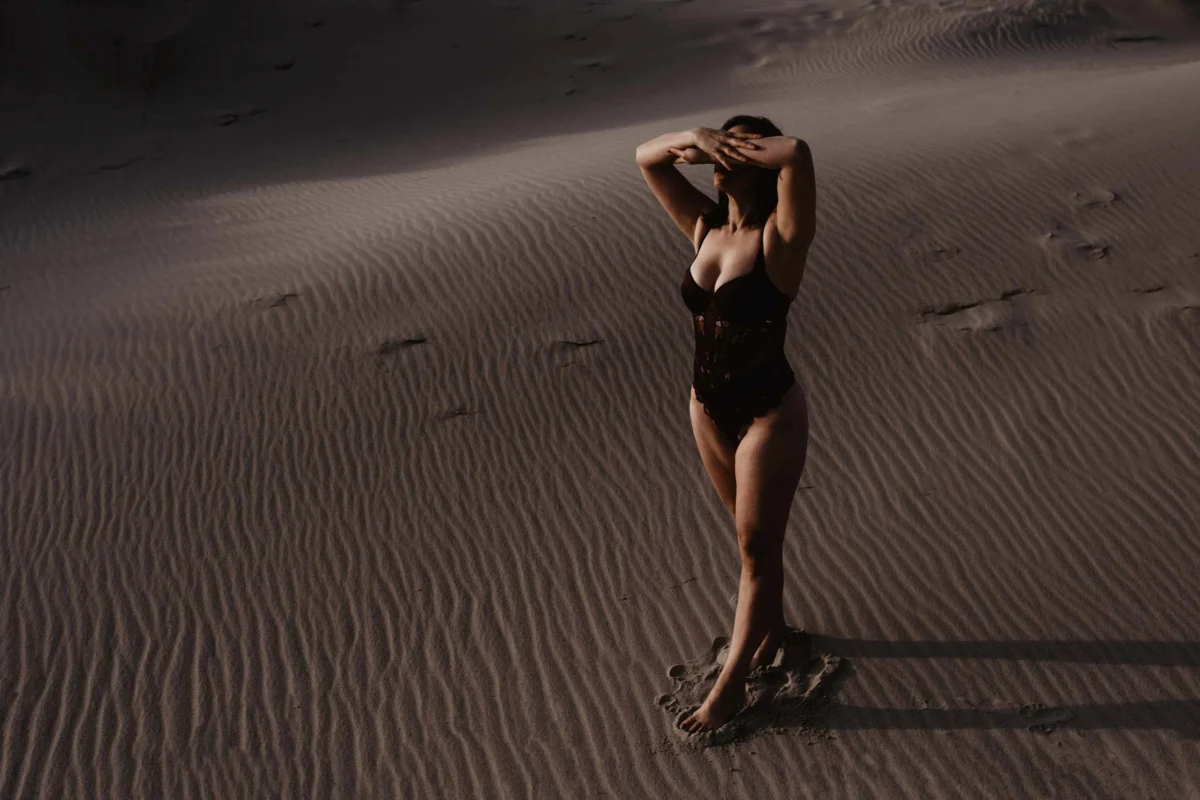Vrouw loopt over het strand in zwart badpak met warm licht van de avondzon, ze heeft haar handen op elegante wijze gedrapeerd over haar gezicht om de zon tegen te houden.