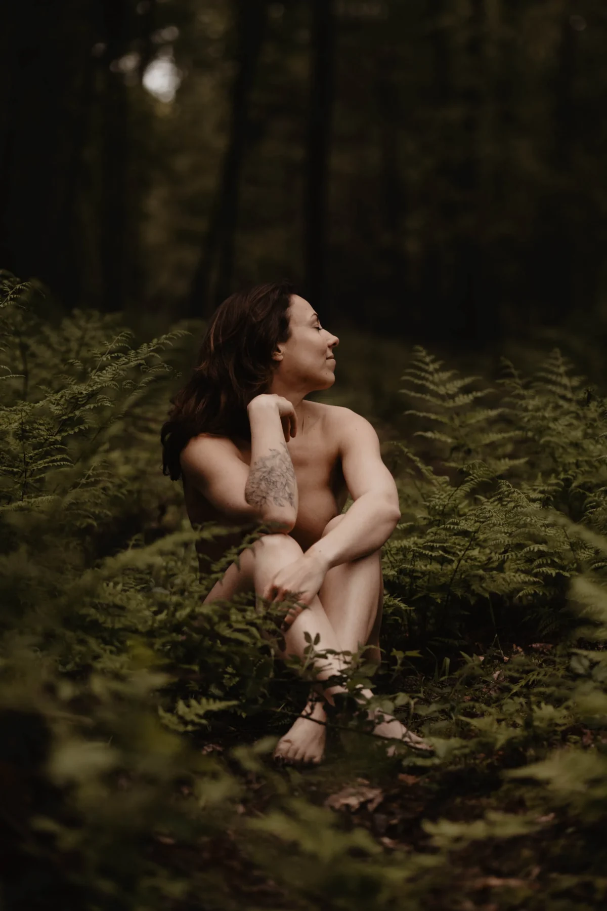 vrouw zit in het bos in een veld met varens, haar witte huid komt mooi uit in het groen van de varens, ze heeft een tattoo op haar onderarm en kijkt weg van de lens.