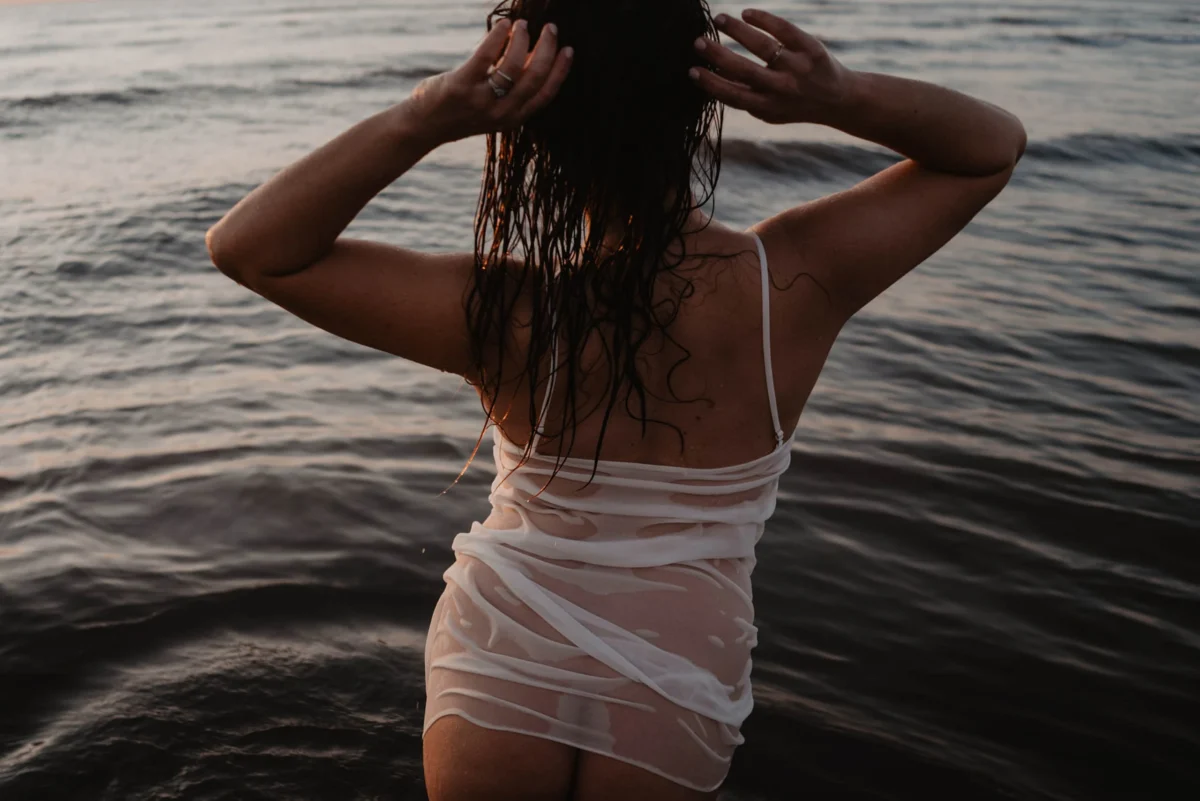 vrouw in nat wit hemd met spaghetti bandjes van achteren te zien, staat in het water met handen omhoog in het natte haar