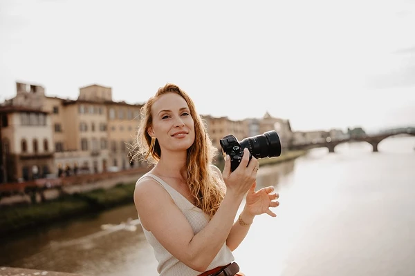Angela de fotograaf van Shapes Shadows met camera in haar hand met een rivier en oude stad uit focus op de achtergrond