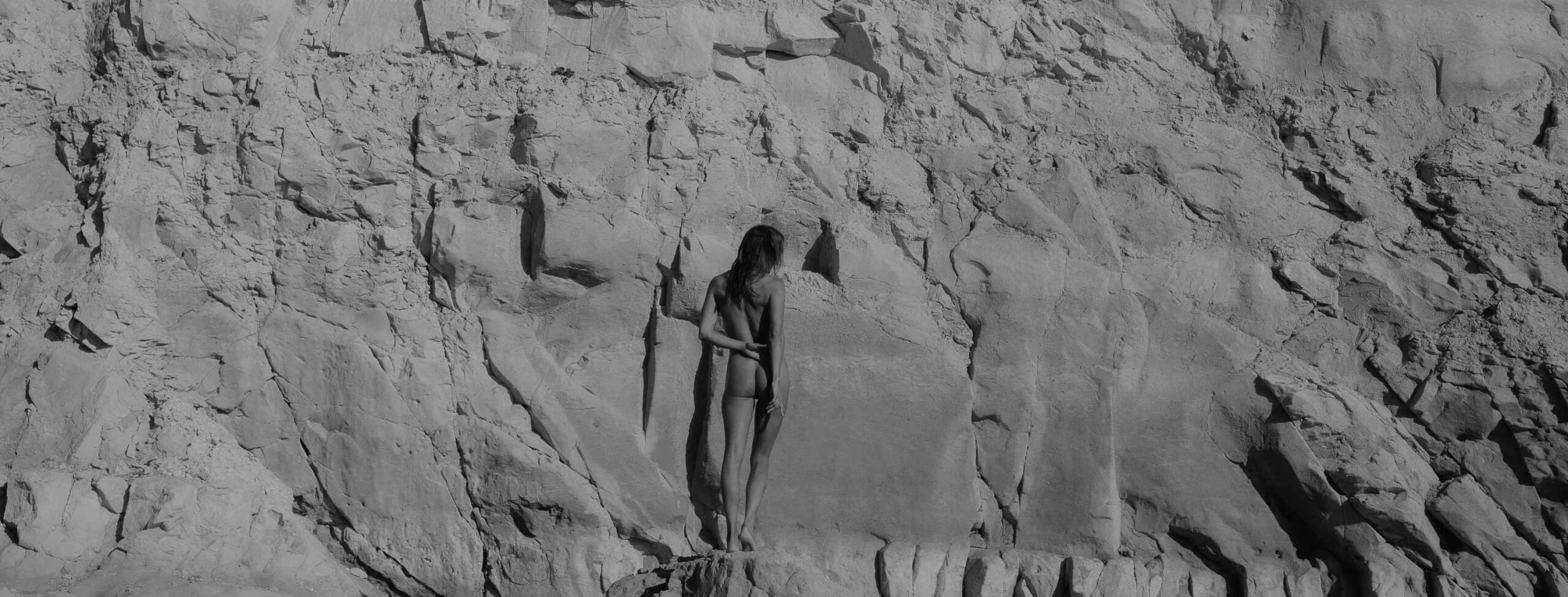 kunstzinnige zwart-wit foto van vrouw naakt van achteren te zien voor een rotspartij, ze blend in met het landschap, haar zachte vormen steken af tegen de ruigheid van de rots. Wat boudoir fotografie is, is hier te zien.