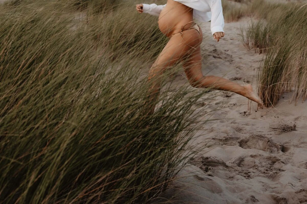 award winnen de foto van zwangere vrouw die door de duinen rent vanaf onderlichaam te zien in beweging met groen helmgras en buik artistiek in harmonie.