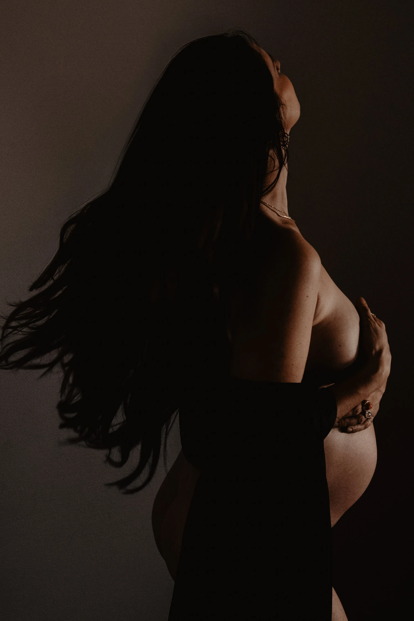 elegante foto met sepia effect van zwangere vrouw die uit de schaduw voorkomt door een lichtbron die op haar buik gericht is. Haar gezicht is naar achteren gebogen en in schaduw gehuld.