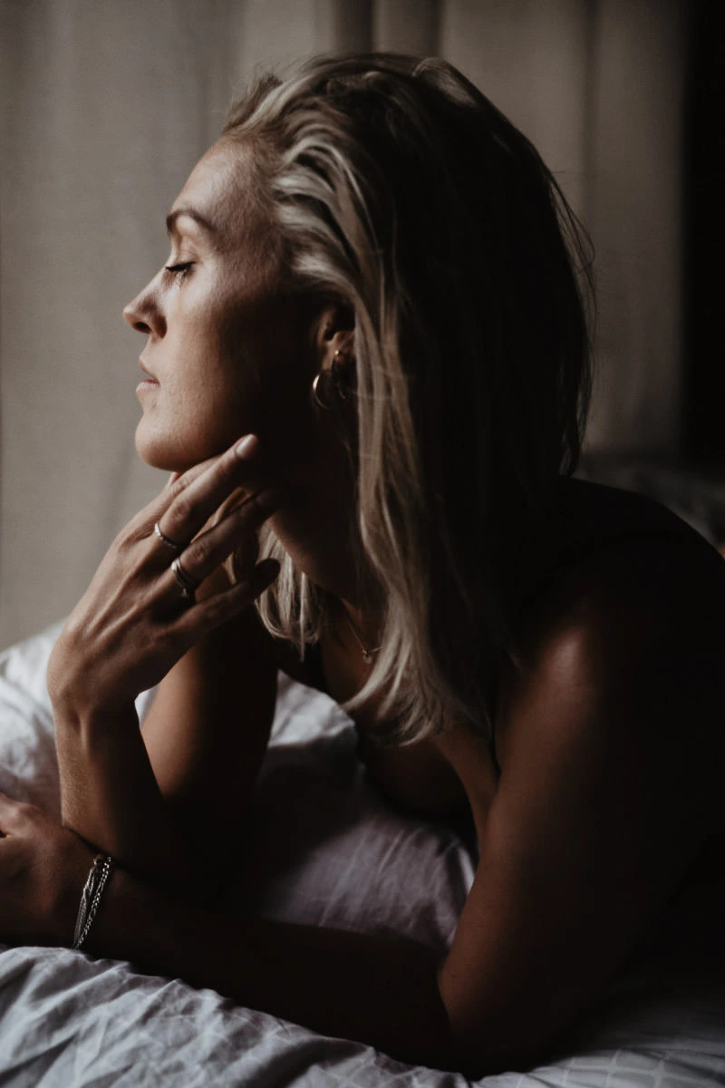 vrouw staat centraal in boudoir portret waarbij zij zacht op haar hand leunt en de focus licht op een zachte lichtbron die de glans van de huid van haar gezicht doet opgloeien en de nadruk legt op haar kaaklijn. Shapes Shadows vindt jouw kracht in fotografie.