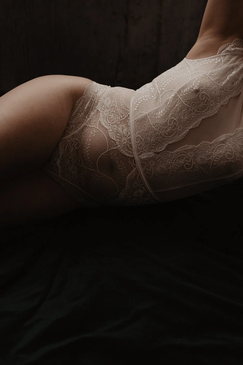 vrouwenlichaam, buik en ronding bovenbeen te zien tegen een donkere achtergrond komt de kleur van de huid door het witte kant heen in een artistiek en zachte vorm.